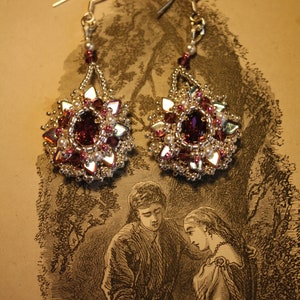 May include: A pair of ornate dangle earrings. Each earring features a large, deep red central stone surrounded by smaller red, pink, and clear crystals, and white pearls. The earrings are set in a silver-toned metal with a beaded design.