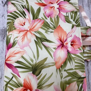 May include: A floral fabric item featuring a tropical orchid design in shades of pink and orange, set against a white background with green leaves. The item has a pink ribbon attached.