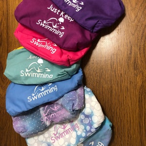 May include: A stack of eight colorful fabric pouches with the phrase "Just Keep Swimming" printed on them. The pouches are in various shades of purple, pink, blue, and green.
