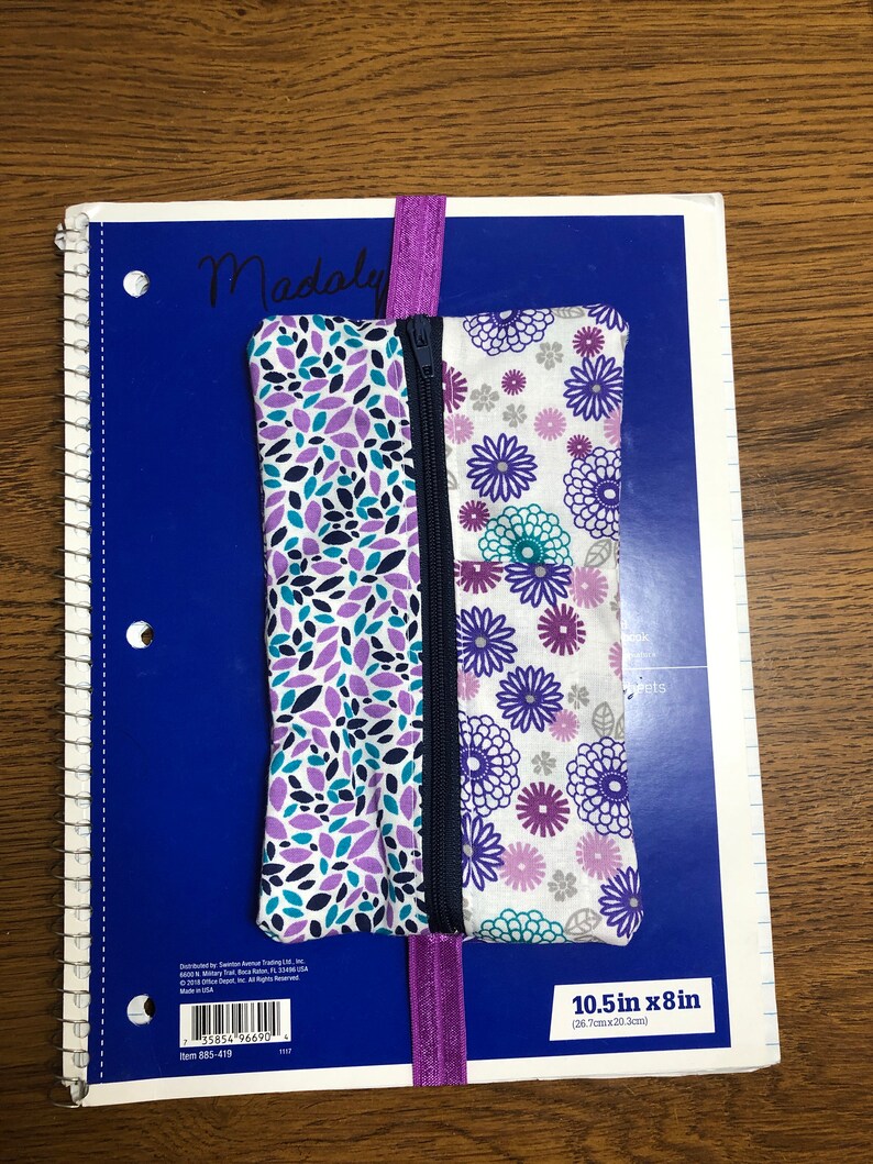 Op de afbeelding: Een ritsvak met een paarse en witte bloemenprint en een paarse elastiek. De pouch is 10,5 inch bij 8 inch (26,7 cm x 20,3 cm).