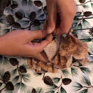 Op de afbeelding: Een paar handen zijn te zien die een bruine en beige stoffen krans vasthouden met een gingerbread man-patroon. De krans wordt op een draadframe geplaatst. De achtergrond is een witte stof met een dennenboom- en dennenappelpatroon.