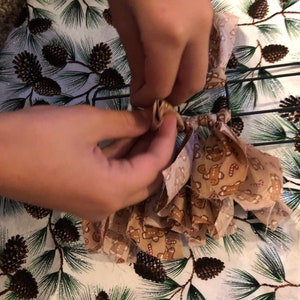 Op de afbeelding: Een paar handen zijn te zien die een bruine strik van stof met een gingerbread man-patroon aan een draadkrans vastbinden.