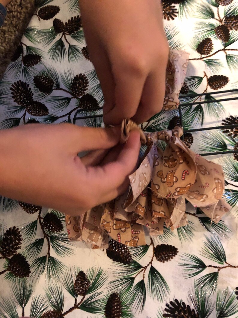 Op de afbeelding: Een paar handen zijn te zien die een bruine strik van stof met een gingerbread man-patroon aan een draadkrans vastbinden.