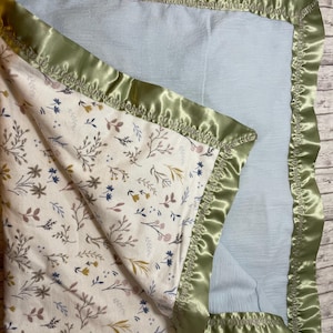 May include: A baby blanket with a floral pattern on a cream background, featuring blue, brown, and yellow accents. The blanket has a light blue interior and is trimmed with a sage green satin border.