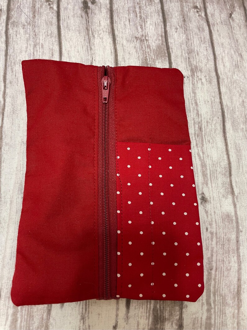 May include: A red zippered pouch with a white polka dot fabric panel on one side. The pouch is made of a soft, red fabric.
