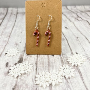 May include: A pair of silver dangle earrings with red and white candy cane charms. The earrings are displayed on a brown card.