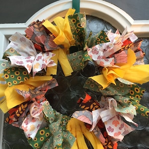Op de afbeelding: Een kleurrijke stoffen krans met gele, groene, witte en bruine stoffen restjes. De krans is versierd met zonnebloem- en bladmotieven.