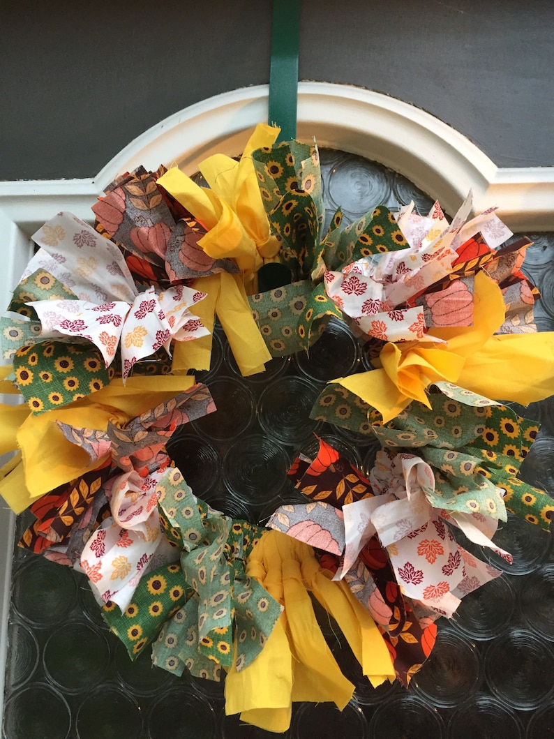 Op de afbeelding: Een kleurrijke stoffen krans met gele, groene, witte en bruine stoffen restjes. De krans is versierd met zonnebloem- en bladmotieven.