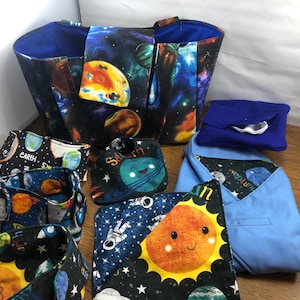 May include: A blue tote bag with a space-themed print featuring planets, stars, and the words "Earth" and "Saturn". The bag is filled with smaller fabric squares with similar prints, including a sun with a smiling face.