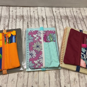 Op de afbeelding: Drie notitieboekjes met ritsvakken en pennenhouders. De notitieboekjes hebben verschillende bloemenpatronen: blauw en geel, roze en wit en rood en wit. De notitieboekjes liggen op een houten oppervlak.