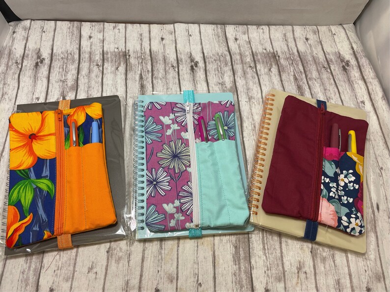Op de afbeelding: Drie notitieboekjes met ritsvakken en pennenhouders. De notitieboekjes hebben verschillende bloemenpatronen: blauw en geel, roze en wit en rood en wit. De notitieboekjes liggen op een houten oppervlak.