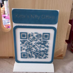 May include: A white and teal QR code display stand. The teal top section reads "Katie's Nifty Giffies" in embossed lettering. The stand features a QR code in the center, with a teal border.