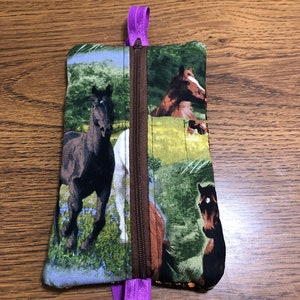Op de afbeelding: Een klein ritsvakje met een paarse rits. Het vakje is gemaakt van stof met een paardenprint. De paarden zijn bruin, zwart en wit en staan tegen een groene achtergrond.