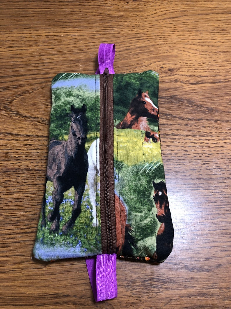 Op de afbeelding: Een klein ritsvakje met een paarse rits. Het vakje is gemaakt van stof met een paardenprint. De paarden zijn bruin, zwart en wit en staan tegen een groene achtergrond.