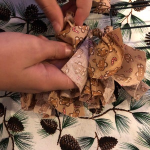 Op de afbeelding: Een paar handen zijn te zien die een bruine strik van stof met een gingerbread man-patroon vasthouden. De strik wordt om een ​​draadkrans gebonden.