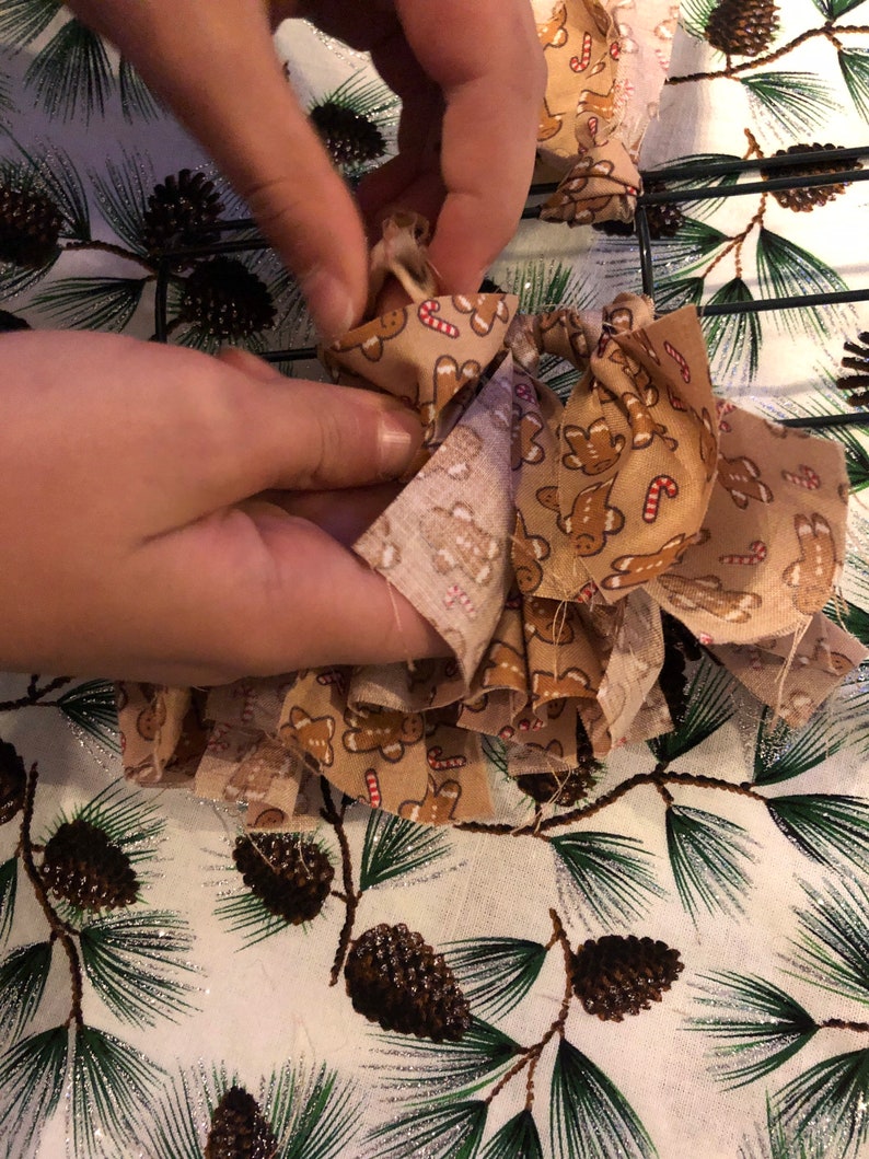 Op de afbeelding: Een paar handen zijn te zien die een bruine strik van stof met een gingerbread man-patroon vasthouden. De strik wordt om een ​​draadkrans gebonden.