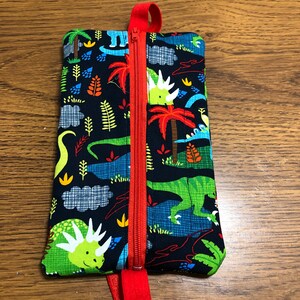 Op de afbeelding: Een klein ritsvakje met een rode rits en een rode band. Het vakje is gemaakt van stof met een dinosaurusprint in tinten groen, blauw en oranje op een zwarte achtergrond.