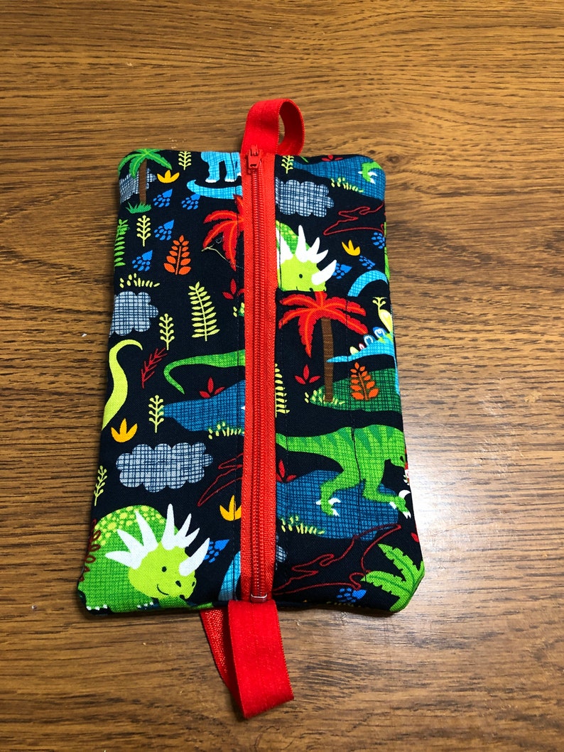 Op de afbeelding: Een klein ritsvakje met een rode rits en een rode band. Het vakje is gemaakt van stof met een dinosaurusprint in tinten groen, blauw en oranje op een zwarte achtergrond.