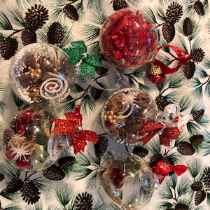 May include: Five clear glass Christmas ornaments filled with various decorative items. The ornaments include red and white confetti, gold and brown pine cones, white beads, and a red bow.