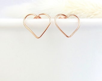 Puede incluir: Un par de pendientes de botón en forma de corazón de oro rosa. Los pendientes tienen un diseño minimalista. La forma de corazón es abierta, formada por un alambre fino. Los pendientes se muestran sobre una superficie blanca.