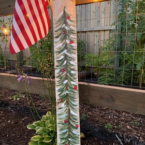 May include: A wooden sign painted to look like a Christmas tree with red baubles. The sign is standing in a garden with green plants and brown mulch.