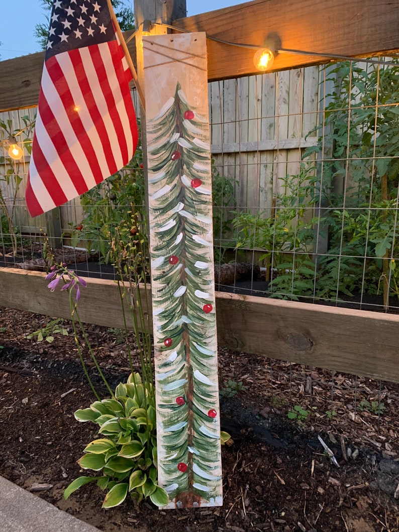 May include: A wooden sign painted to look like a Christmas tree with red baubles. The sign is standing in a garden with green plants and brown mulch.