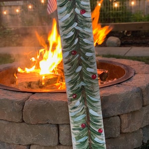 May include: A hand-painted wooden sign featuring a green Christmas tree design with red baubles. The sign is positioned in front of a fire pit with flames.