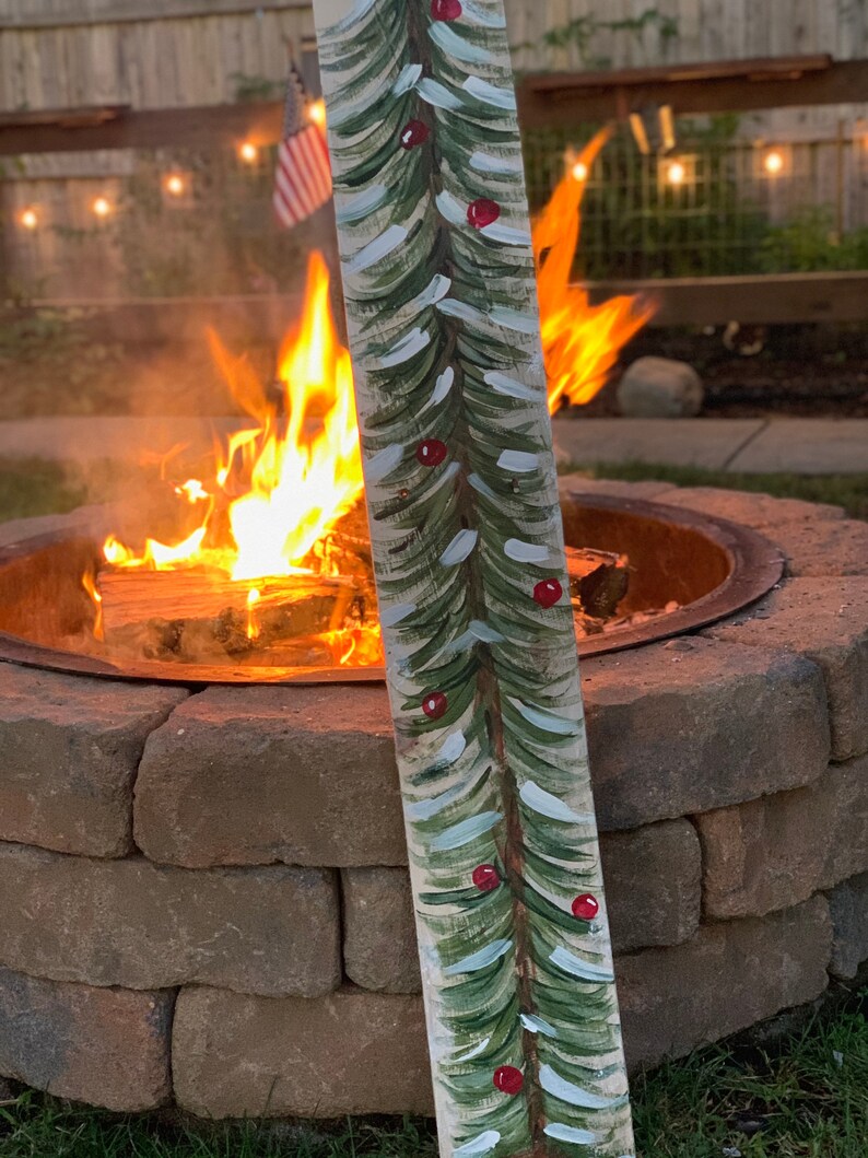 May include: A hand-painted wooden sign featuring a green Christmas tree design with red baubles. The sign is positioned in front of a fire pit with flames.