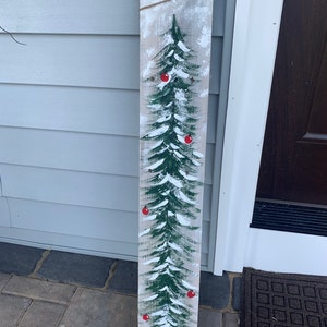 May include: A wooden sign featuring a painted green Christmas tree with red baubles and white snow. The sign has a distressed, rustic appearance.