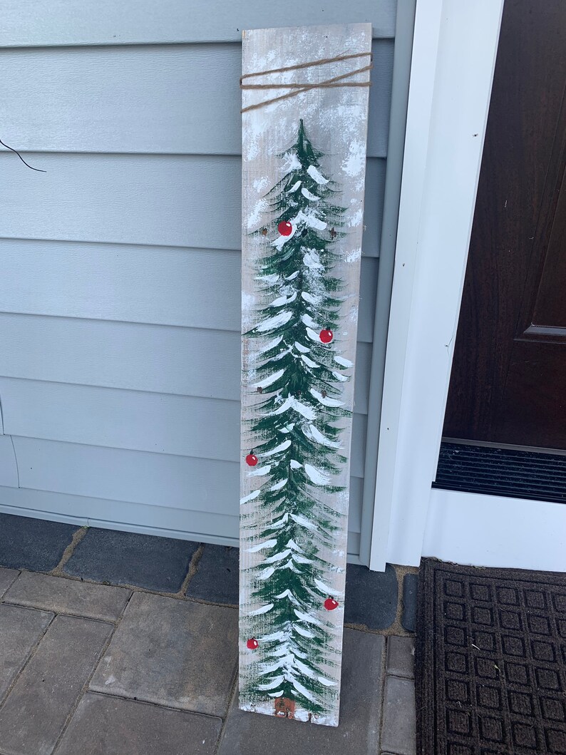May include: A wooden sign featuring a painted green Christmas tree with red baubles and white snow. The sign has a distressed, rustic appearance.