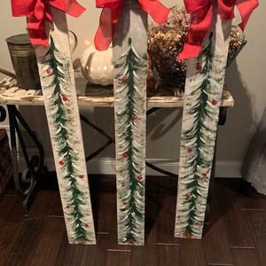 May include: Three wooden planks painted to look like Christmas trees with red bows and red berries. The planks are standing upright on a wooden floor.