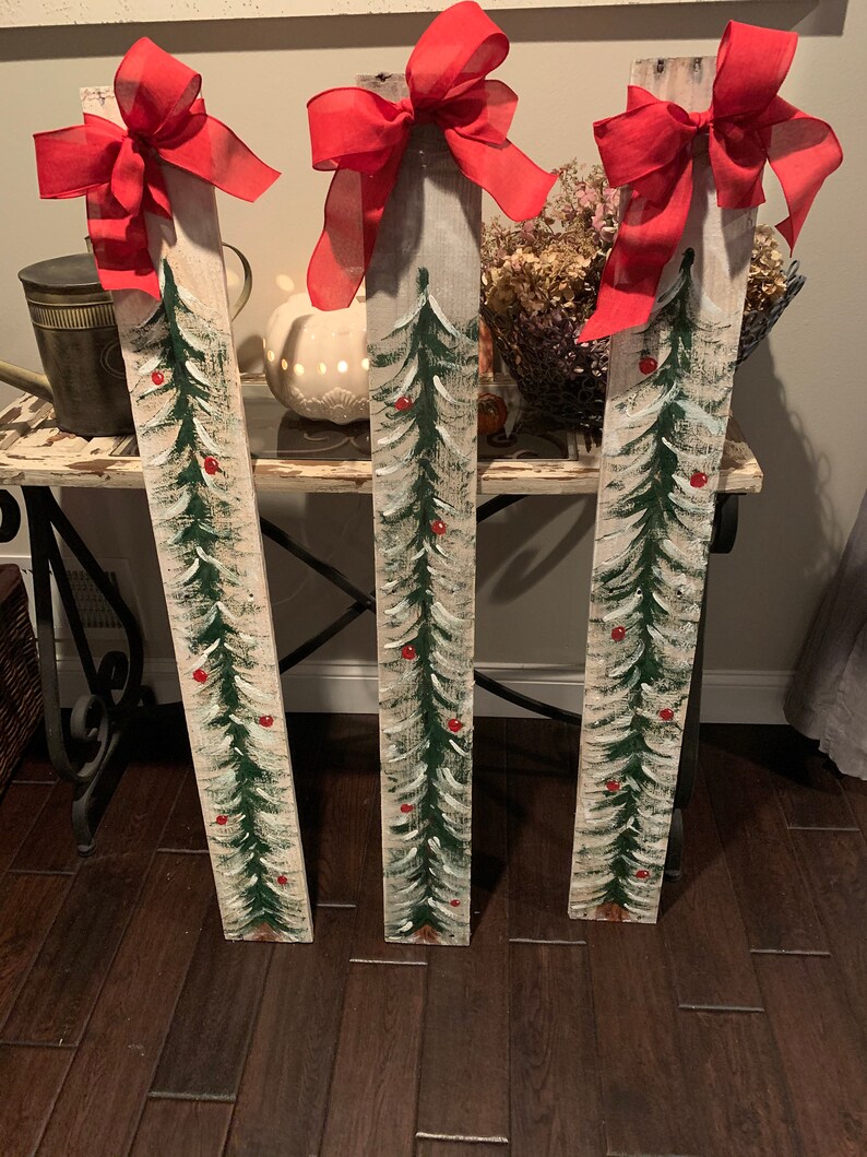 May include: Three wooden planks painted to look like Christmas trees with red bows and red berries. The planks are standing upright on a wooden floor.