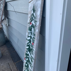 May include: A rustic wooden sign featuring a painted green Christmas tree with red baubles. The sign is tied with twine and has a whitewashed background.