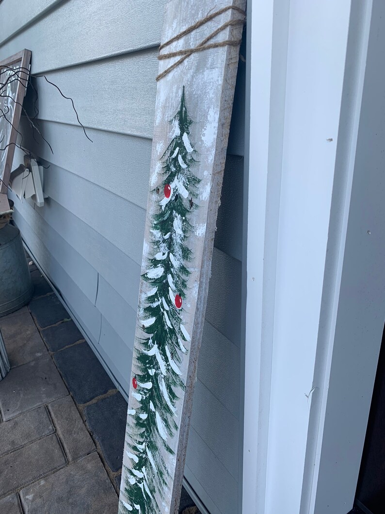 May include: A rustic wooden sign featuring a painted green Christmas tree with red baubles. The sign is tied with twine and has a whitewashed background.