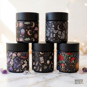 May include: Five cylindrical black jars with matte black lids, each featuring a unique colorful pattern on a black background, including celestial motifs, botanical illustrations, tarot cards, rabbits, mushrooms, and roses, arranged in a group.