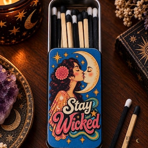 May include: A rectangular metal tin filled with wooden matches featuring black and white tips. The lid displays a colorful illustration of a person kissing a crescent moon, surrounded by stars, with the text Stay Wicked in stylized lettering.