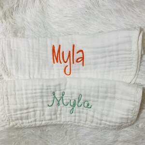 May include: Two white muslin burp cloths with the name "Myla" embroidered on them. The top cloth has the name embroidered in orange thread, and the bottom cloth has the name embroidered in green thread.