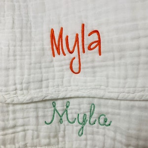 May include: A white muslin baby blanket with the name "Myla" embroidered in orange thread and "Myla" embroidered in green thread.