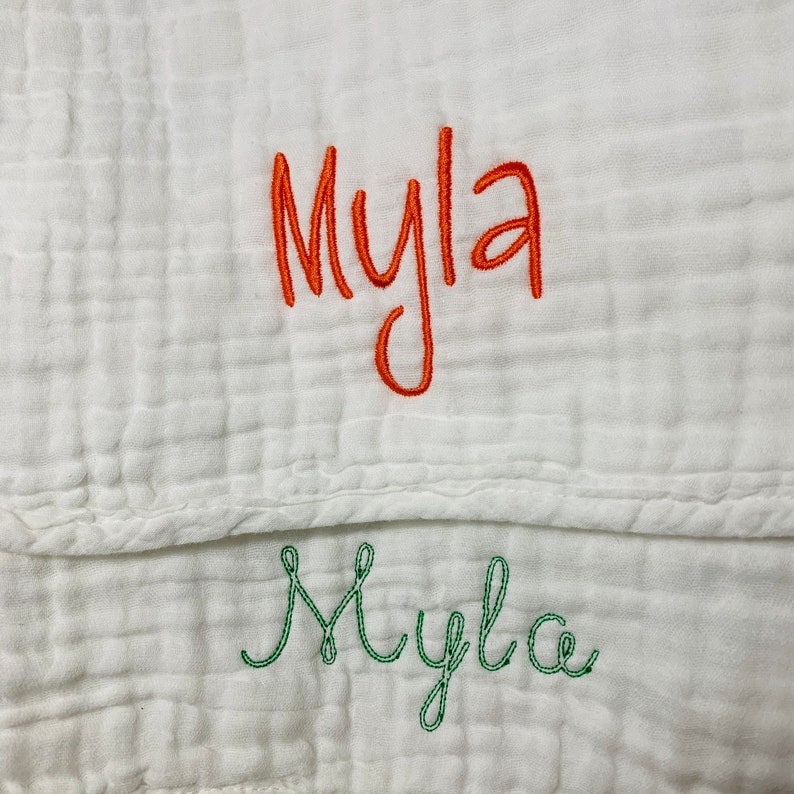 May include: A white muslin baby blanket with the name "Myla" embroidered in orange thread and "Myla" embroidered in green thread.