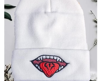 May include: White beanie featuring an embroidered design of a mouth with sharp teeth and a red tongue. The beanie is made of a soft, knitted material. The mouth design is outlined in black and red.