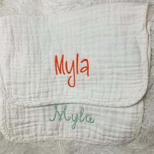 May include: White muslin baby burp cloth with the name "Myla" embroidered in orange and green thread.