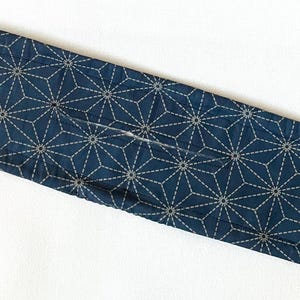 May include: A navy blue fabric hair accessory with a geometric starburst pattern in white stitching. The accessory is long and oval-shaped, likely a hair wrap or bun maker, and rests on a white surface.