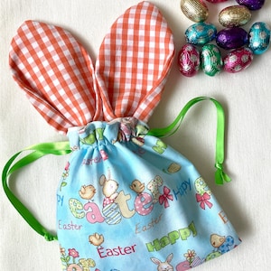May include: A blue Easter-themed drawstring bag with bunny ears and green ribbon ties. The bag is decorated with the word "Easter" and illustrations of bunnies, chicks, and eggs. Several foil-wrapped chocolate eggs in different colours are also visible.