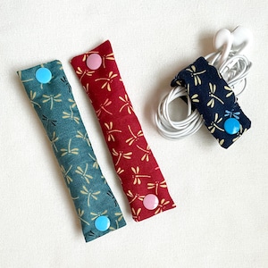 May include: Three fabric earbud organizers in teal, red, and navy blue, each with a dragonfly pattern and a snap closure. One organizer holds white earbuds. The teal and red organizers are side by side. The navy blue organizer is wrapped around the earbuds.
