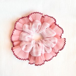 May include: A pink scrunchie with a pleated design and a ruffled edge. The scrunchie has a light pink and white plaid center. The edge is trimmed with a dark burgundy color.