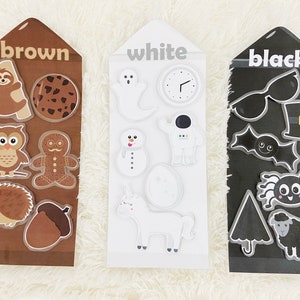 May include: Three cards with different colors, brown, white, and black, each with various images on them. The brown card has a sloth, a cookie, an owl, a gingerbread man, a hedgehog, and an acorn. The white card has a ghost, a clock, a snowman, an astronaut, an egg, and a unicorn. The black card has sunglasses, a top hat, a bat, a spider, an umbrella, and a sheep.