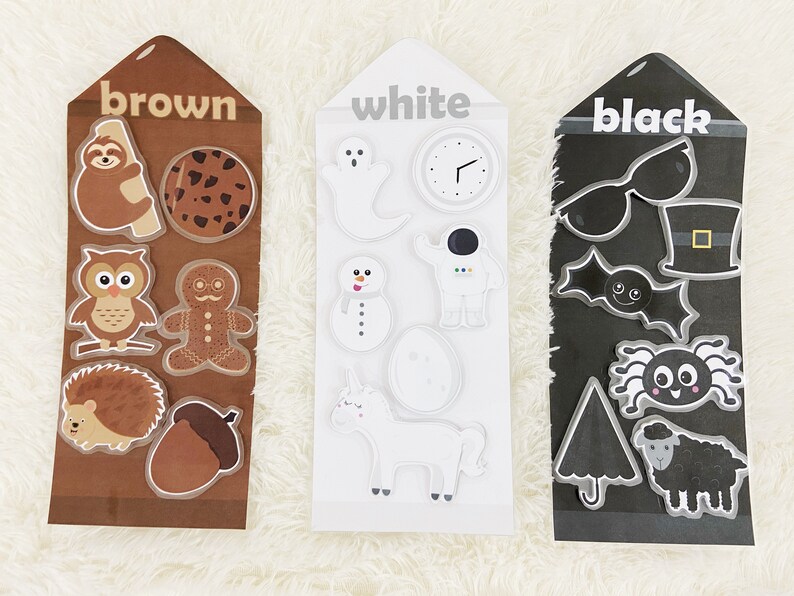 May include: Three cards with different colors, brown, white, and black, each with various images on them. The brown card has a sloth, a cookie, an owl, a gingerbread man, a hedgehog, and an acorn. The white card has a ghost, a clock, a snowman, an astronaut, an egg, and a unicorn. The black card has sunglasses, a top hat, a bat, a spider, an umbrella, and a sheep.
