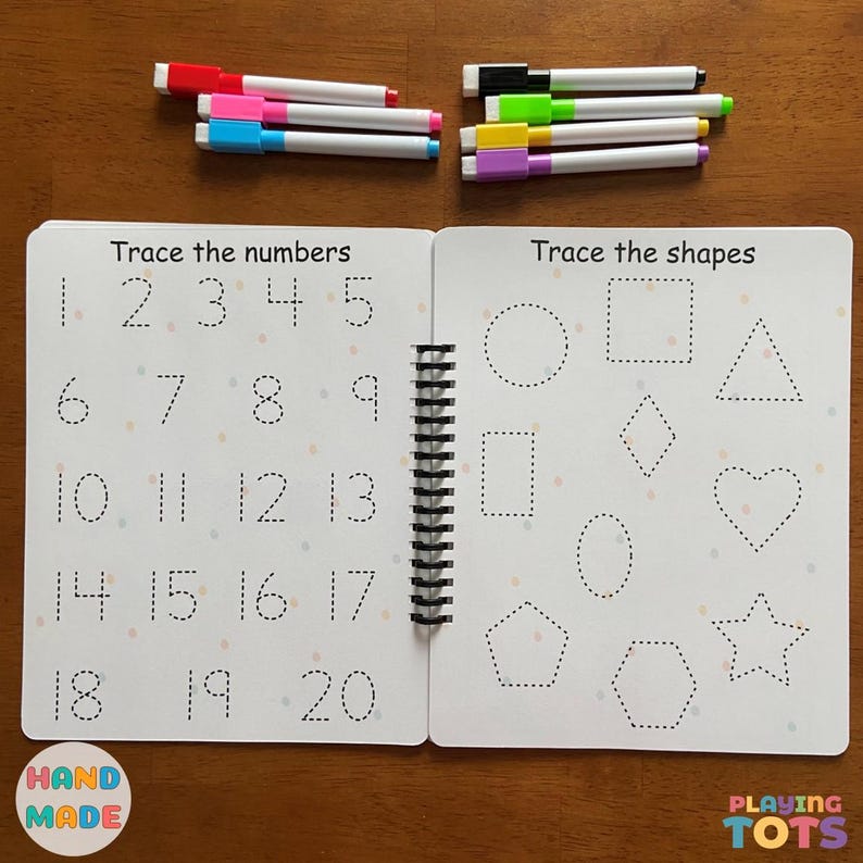 May include: A spiral-bound learning book with the text "Trace the numbers" and "Trace the shapes" on white pages. The book includes dotted lines for tracing numbers and shapes. Several colorful markers are arranged around the book.
