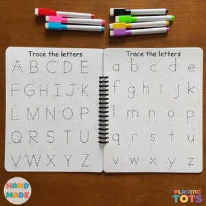 May include: A spiral-bound tracing book with uppercase and lowercase letters, and colorful markers. The book is open, displaying dotted letters for tracing. The words "Trace the letters" are at the top of each page. The book is labeled "Hand Made" and "Playing Tots".