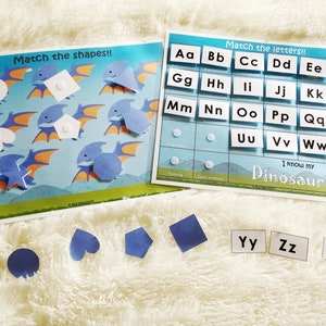 Puede incluir: Un juego de tarjetas educativas para ni&ntilde;os con dinosaurios y letras del alfabeto. Las tarjetas son azules y blancas con ilustraciones coloridas de dinosaurios. Las letras est&aacute;n impresas en negro sobre tarjetas blancas. Las tarjetas est&aacute;n dispuestas en una cuadr&iacute;cula sobre una superficie blanca y peluda.